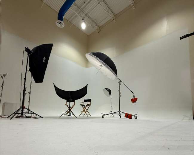 The image shows a photography studio setup with lighting equipment, backdrops, and two empty director's chairs on a white floor.