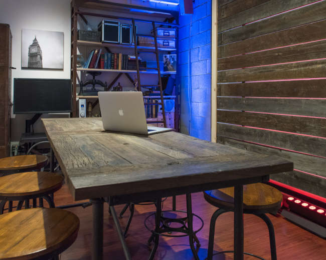 A conference room with a wooden table and four stools, a laptop on the table, and a wall featuring shelves filled with books and decorative items.