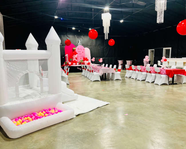 The image shows a party venue decorated with pink balloons, a long dining table set with white chairs, and a white inflatable bounce house filled with colorful balls.