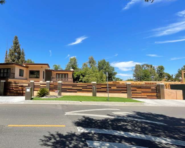A modern house with a wooden fence and landscaped yard is visible from the street under a clear blue sky.