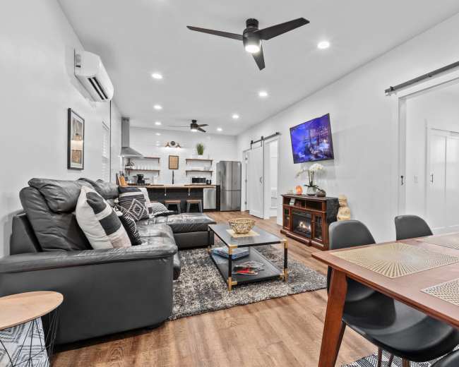 The image shows a modern living space featuring a black leather sofa, a dining area with a wooden table, and an open kitchen with stainless steel appliances.