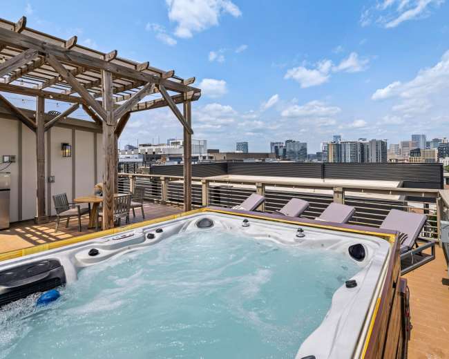 A rooftop deck features a hot tub with city skyline views and lounge chairs under a pergola.