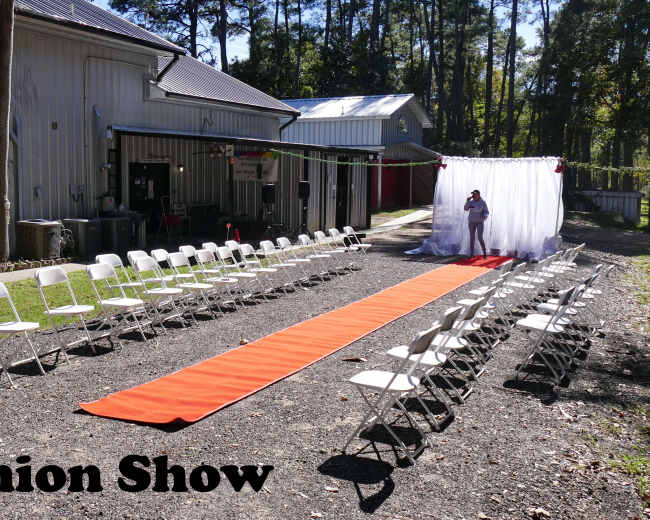 A fashion show setup features an orange runway with white folding chairs arranged on either side, near a backdrop draped in white fabric.