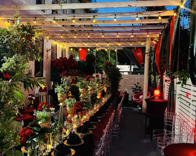 The image shows a beautifully arranged outdoor dining setup under a wooden pergola, adorned with greenery and string lights.
