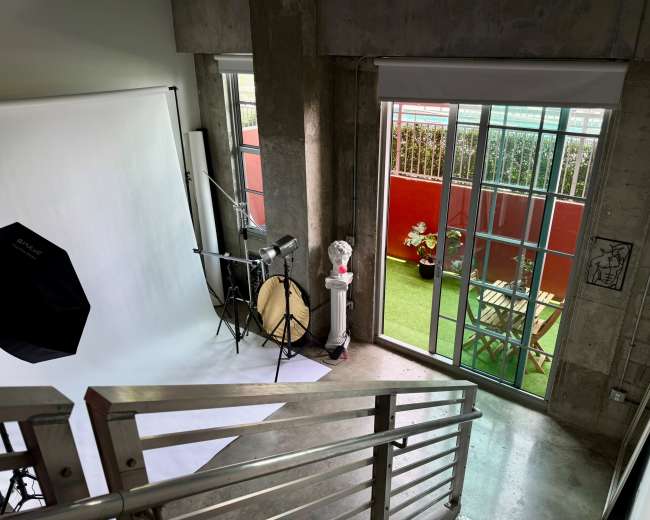 The image shows a photography studio with a white backdrop, lighting equipment, and an adjacent outdoor area visible through large glass doors.