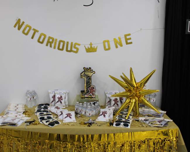 A decorated table displays a gold-themed birthday setup featuring a "Notorious One" banner, a large number one centerpiece, and party favors.