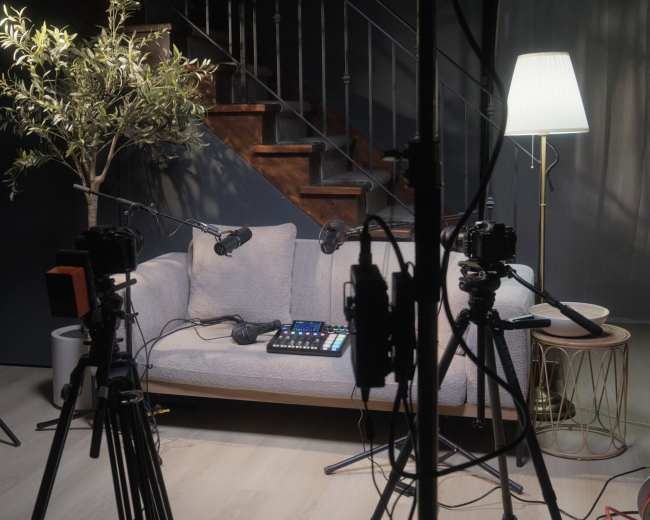 A gray sofa is positioned under a staircase, surrounded by cameras and lighting equipment, with a small table and a plant nearby.
