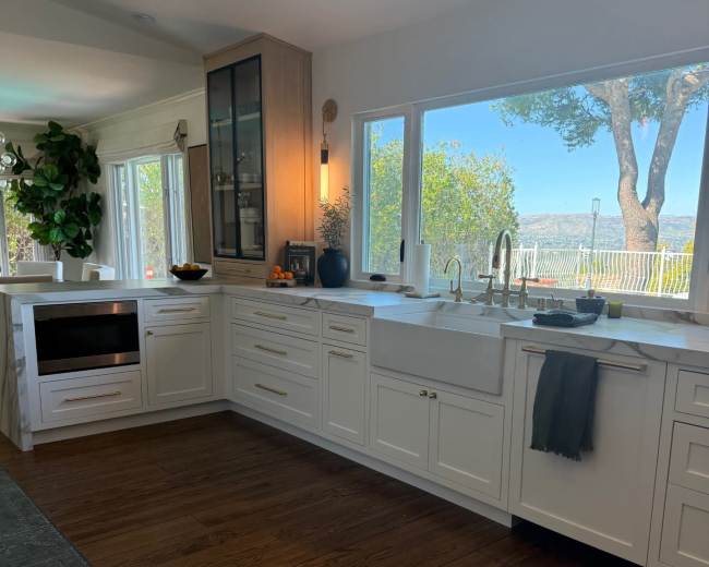The kitchen features white cabinetry, a large window overlooking a scenic view, and wooden flooring.