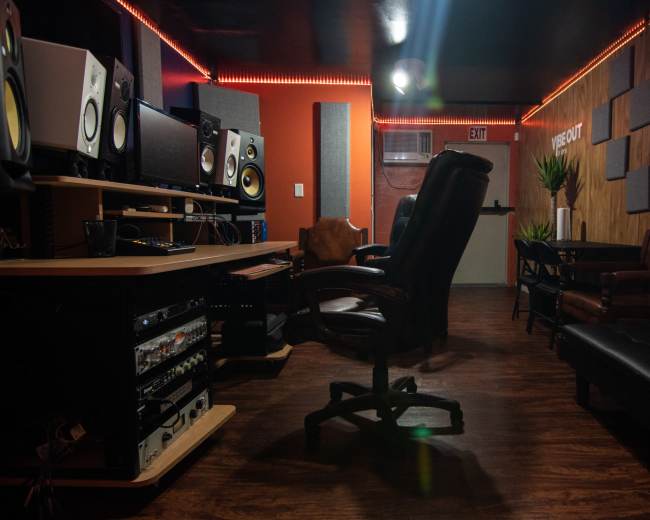 The image shows a music studio with various audio equipment, a comfortable seating area, and warm wooden paneling.