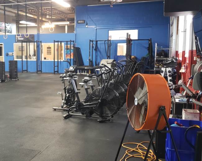 The image shows a spacious gym interior with exercise equipment, including rowing machines and a large fan, set against blue and gray walls.