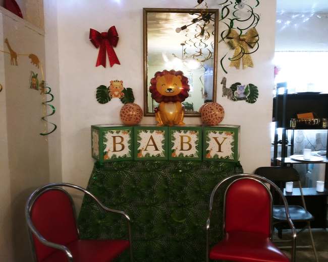 The scene depicts a decorated corner with a mirror, a lion decoration, and blocks spelling "BABY," surrounded by festive adornments like ribbons and leaves.