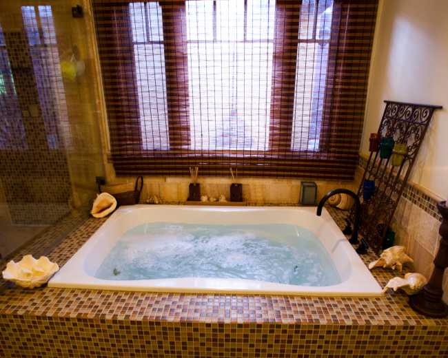 A whirlpool bathtub is situated in a luxurious bathroom featuring mosaic tiled walls and a wooden window with blinds.