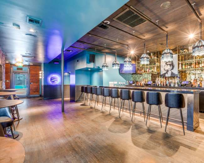 The image shows a modern bar interior with a long, dark countertop and stylish pendant lighting, featuring shelves lined with various bottles in the background.