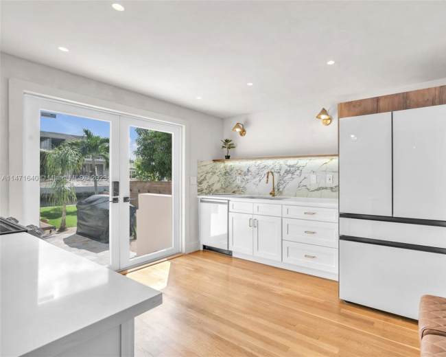 The image shows a modern kitchen with white cabinetry, a large fridge with wooden accents, and a marble backsplash, illuminated by natural light from a nearby door.