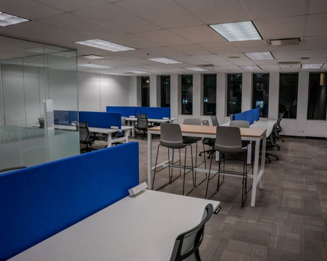 The image shows an office space with several workstations featuring blue partitions and a mix of desk and high-top seating, all illuminated by overhead lights.