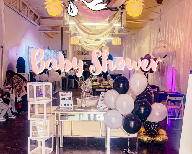 The image shows a beautifully decorated venue for a baby shower, featuring balloons, a welcoming sign, and festive drapery.