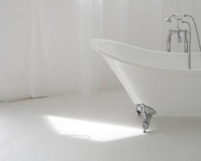 A freestanding white bathtub is positioned near a window with sheer white curtains in a bright, minimalistic room.