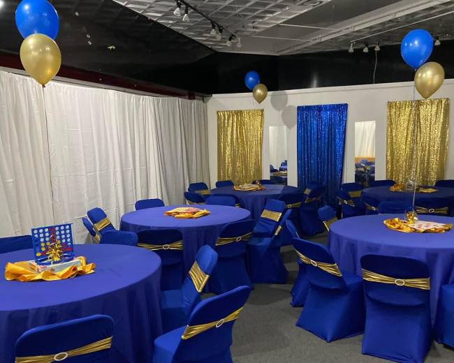 The room is set up for an event with round tables covered in blue tablecloths and decorated with gold balloons and accessories.