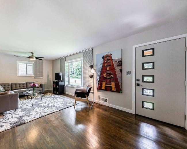 The image shows a modern living room featuring a gray sectional sofa, a round glass coffee table, and a large window that lets in natural light, with a door and a distinctive piece of wall art.