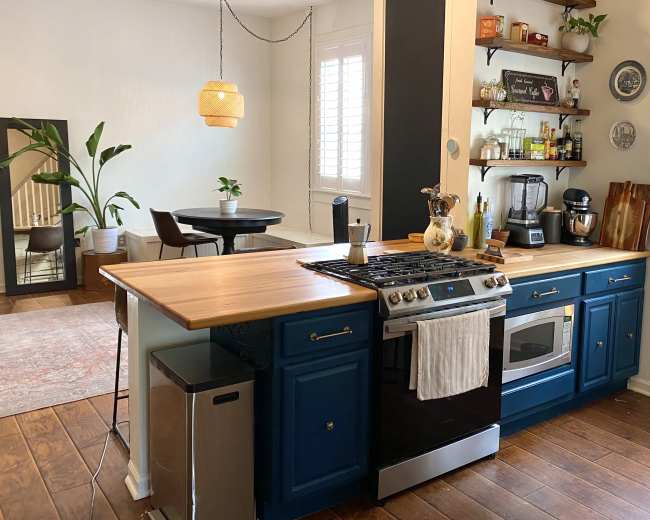 The image depicts a modern kitchen featuring blue cabinets, a gas stove, and a wooden countertop, with an adjoining dining area visible in the background.