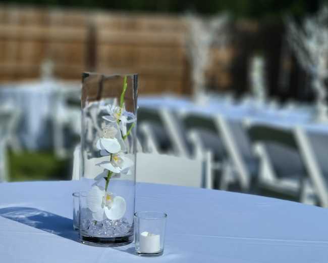 A clear vase with white orchids and small stones is placed on a table set for an event, surrounded by empty chairs in the background.