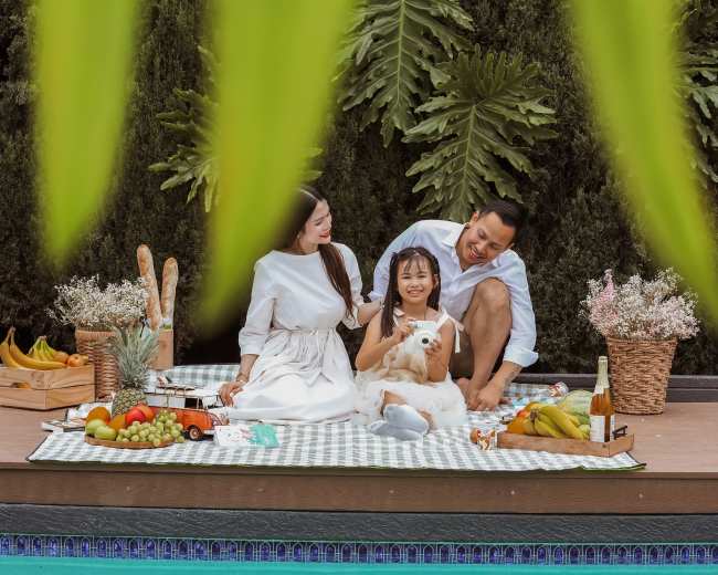 A family enjoys a picnic by a poolside, surrounded by various fruits and a small dog.