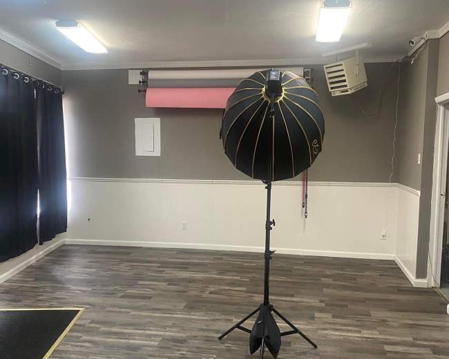 An empty room with wooden flooring, featuring a large photography light umbrella on a stand, dark curtains on one side, and a backdrop holder with a pink backdrop hanging on the wall.