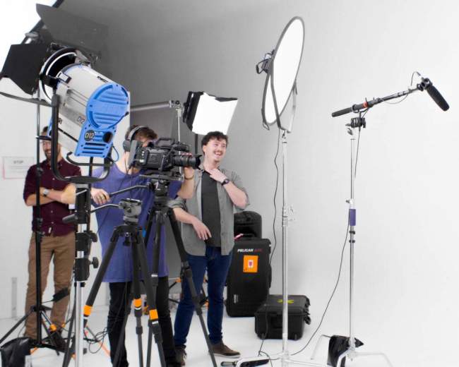 A film crew sets up equipment, including cameras and lights, in a bright studio to shoot a segment featuring a woman.