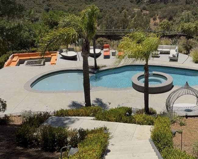 The image shows a landscaped backyard with a swimming pool, palm trees, and a mountainous background.