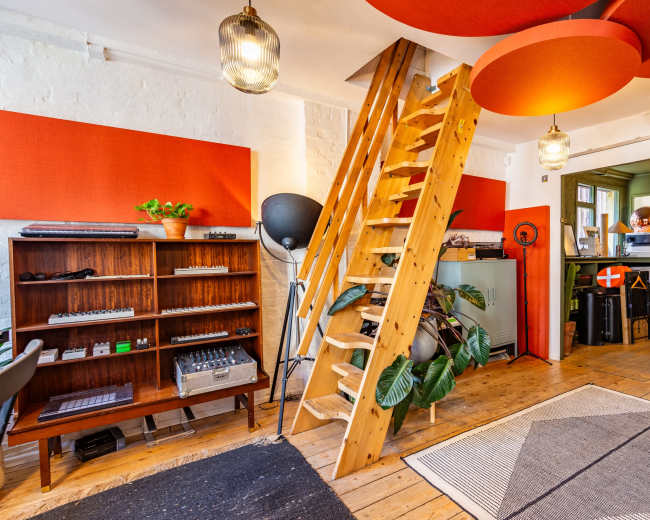 The image showcases a modern interior space featuring wooden stairs leading to a loft area, a bookshelf with various equipment, and vibrant red wall panels.