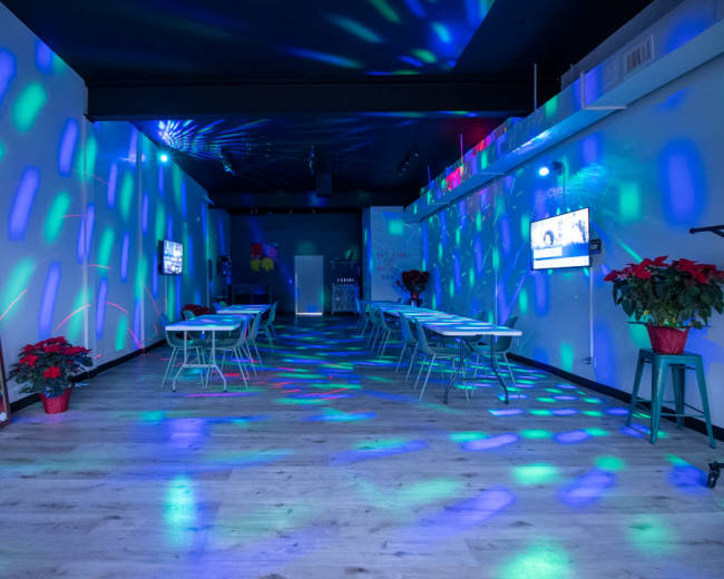 A party venue is illuminated with multicolored lights, featuring decorated tables and a large mirror with a birthday message.
