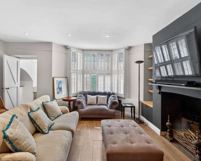 The image shows a cozy living room with a beige sectional sofa, a grey armchair, and a flat-screen TV mounted on a black wall, along with wooden shelves and a fireplace.