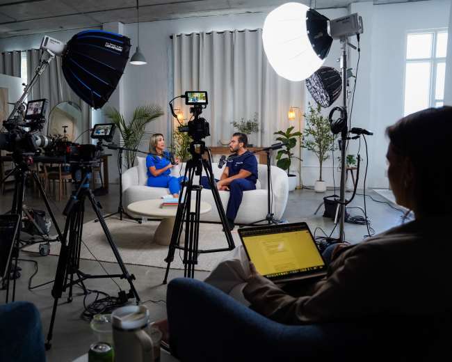 A woman in a blue dress and a man in a dark shirt sit on a couch in a brightly lit studio setup for an interview, surrounded by multiple cameras and lighting equipment.