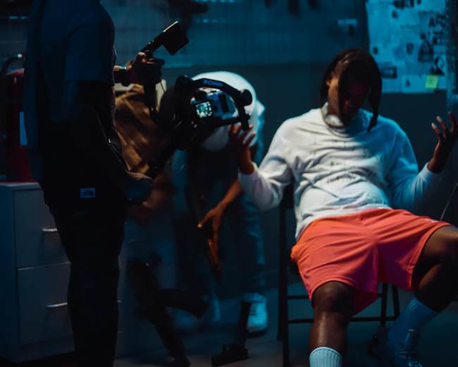 A dimly lit room with three people, one wearing a white hoodie and orange shorts seated in a chair, while another holds a camera and a third appears to be holding a weapon.