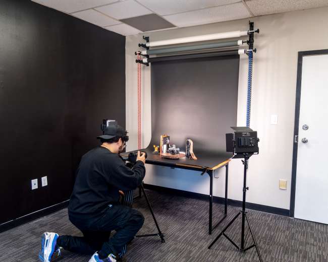 A person is kneeling while taking photographs of various items displayed on a table in a studio setting.