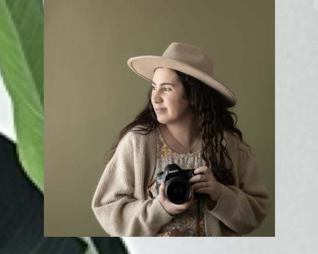 A woman wearing a hat holds a camera while standing in front of a solid green wall.
