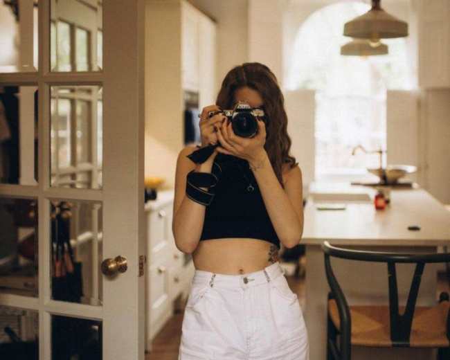 A person stands in a brightly lit kitchen, holding a camera and looking directly at the lens.