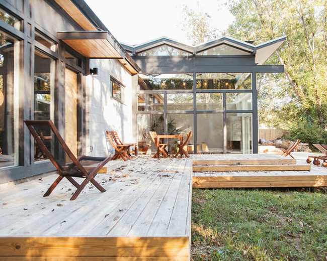 A modern home with large windows features a wooden deck and several outdoor seating areas in a forested setting.