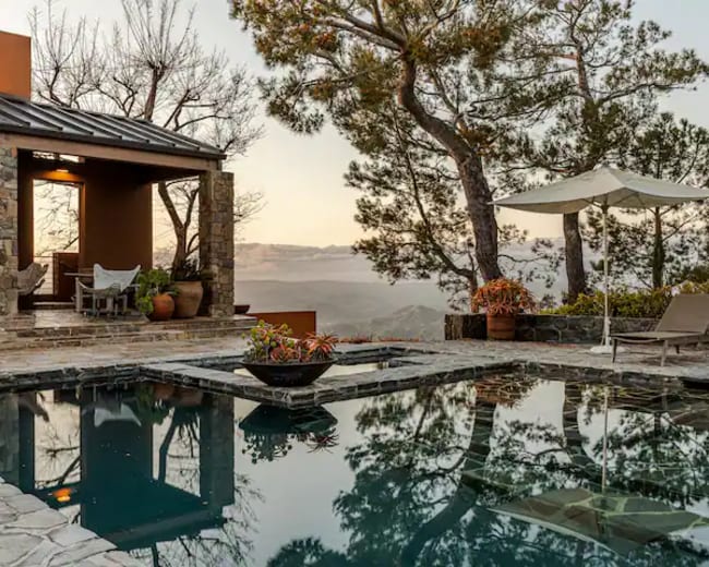 The image shows a modern stone pool area surrounded by trees, with a patio featuring seating and an umbrella.