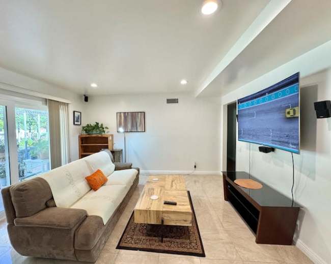 The living room features a sofa, a wooden coffee table, a wall-mounted TV, and sliding glass doors leading to an outdoor area.