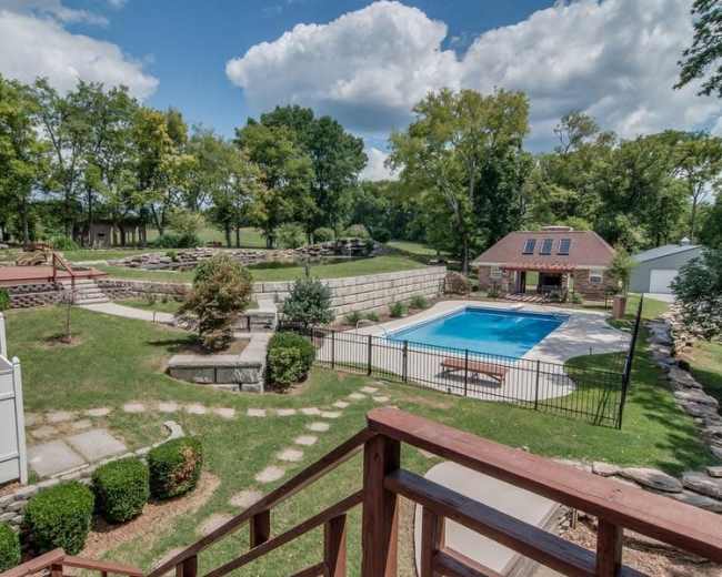 The image shows a landscaped backyard featuring a swimming pool, surrounding trees, a stone wall, and a small building with solar panels.