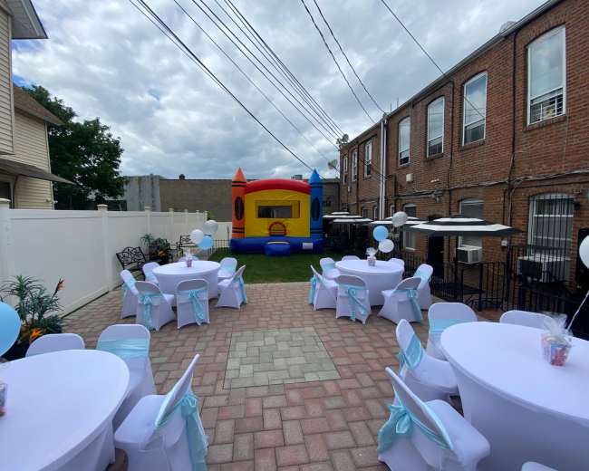 A backyard party setup features round tables with white tablecloths and light blue chair sashes, alongside a colorful inflatable bouncing castle.
