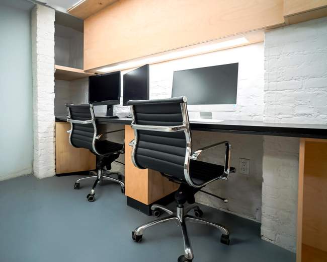 The image shows an office space featuring two modern workstations with black swivel chairs and dual computer monitors on each desk, placed against a brick wall.