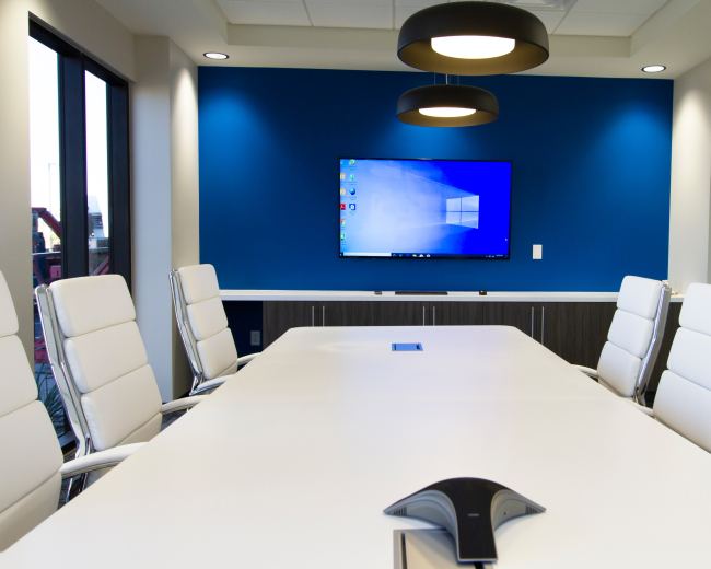 A bright conference room features a long white table, white leather chairs, and a large wall-mounted screen against a blue backdrop.
