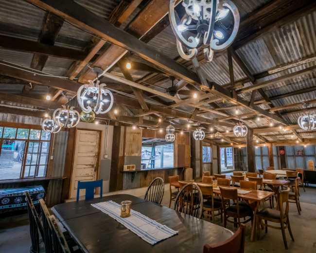 The image shows the interior of a rustic dining area with wooden tables and chairs, industrial-style light fixtures, and exposed beams.