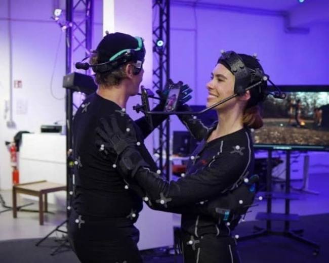 Two people are wearing motion capture suits with sensors while interacting in a studio environment equipped with a display screen.