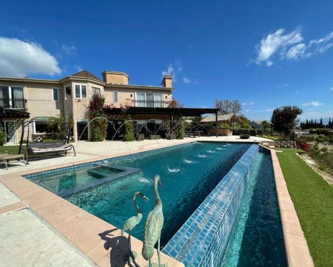 The image shows a swimming pool with decorative fountains set in a backyard next to a house, surrounded by landscaped areas and clear blue skies.