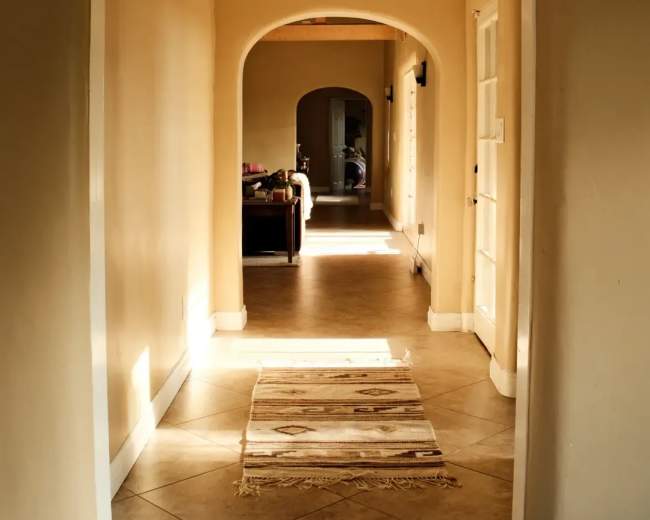 The image shows a long hallway with a patterned rug on the tiled floor, leading to an arched opening and a glimpse of a living area at the end.
