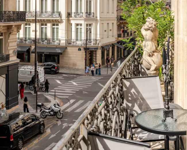 The image shows a balcony overlooking a busy Parisian street, with classic buildings and vehicles visible below.