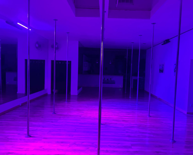 The image shows a dance studio with several pole dance poles illuminated by purple lights, casting a reflective glow on the wooden floor.